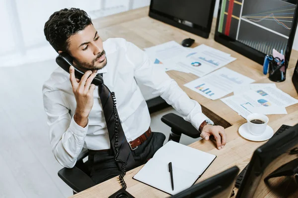 Vue grand angle du commerçant bi-racial parlant au téléphone et assis près de l'ordinateur avec des graphiques — Photo de stock