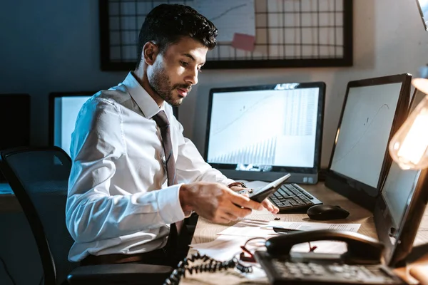 Comerciante bi-racial usando calculadora e sentado perto de computadores com gráficos — Fotografia de Stock