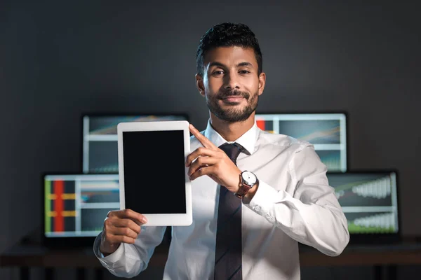 Comerciante bi-racial sonriente sosteniendo tableta digital y computadoras con gráficos en el fondo - foto de stock