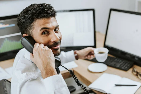 Messa a fuoco selettiva di sorridente trader bi-razziale parlando su smartphone e tenendo in mano tazza — Foto stock