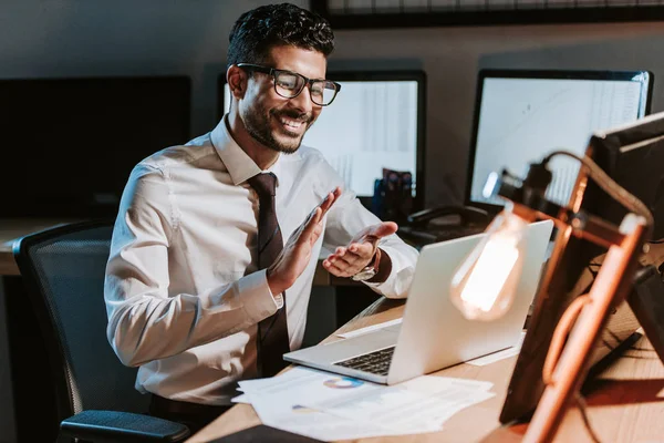 Sonriente y guapo bi-racial comerciante aplaudiendo y mirando portátil - foto de stock