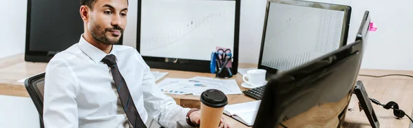 Panoramic shot of bi-racial trader looking at computer and holding paper cup — Stock Photo