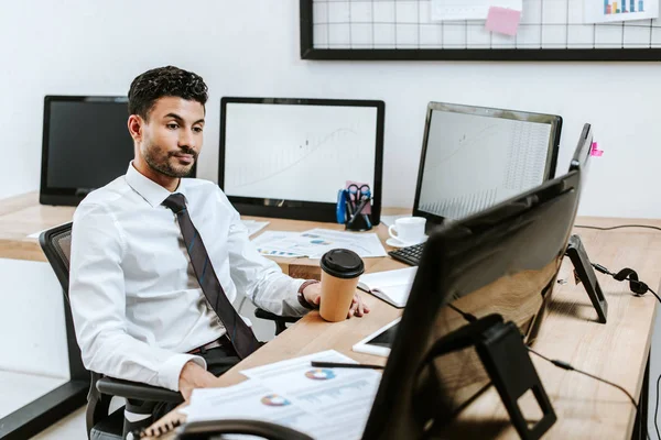 Händler blickt auf Computer und hält Pappbecher in der Hand — Stockfoto