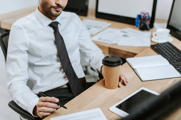 Vista recortada del comerciante bi-racial sosteniendo taza de papel y pluma — Stock Photo