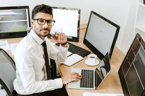 Hochwinkelaufnahme eines lächelnden bi-rassischen Händlers, der neben Computern sitzt und in die Kamera blickt — Stock Photo