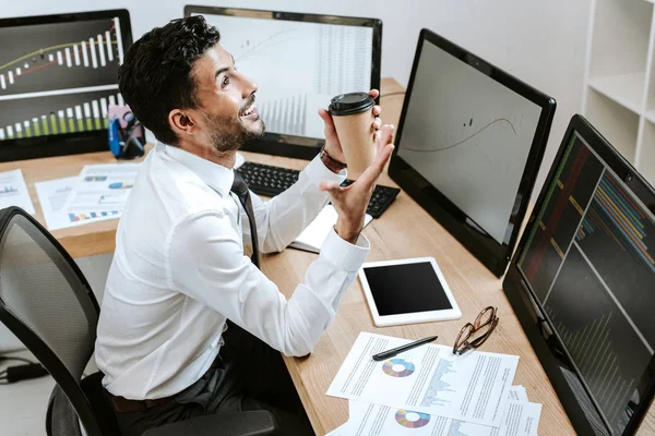 Vista de ángulo alto del comerciante bi-racial sonriente sosteniendo taza de papel y sentado cerca de computadoras con gráficos - foto de stock