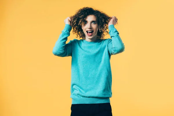 Lockiger Teenager mit offenem Mund, der lockiges Haar berührt, isoliert auf gelb — Stockfoto