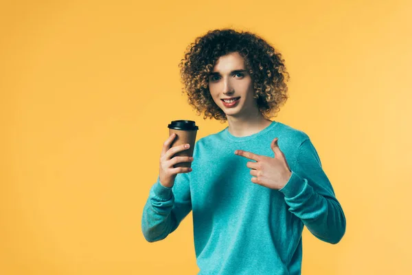 Smiling curly teenager pointing at coffee in paper cup isolated on yellow — Stock Photo