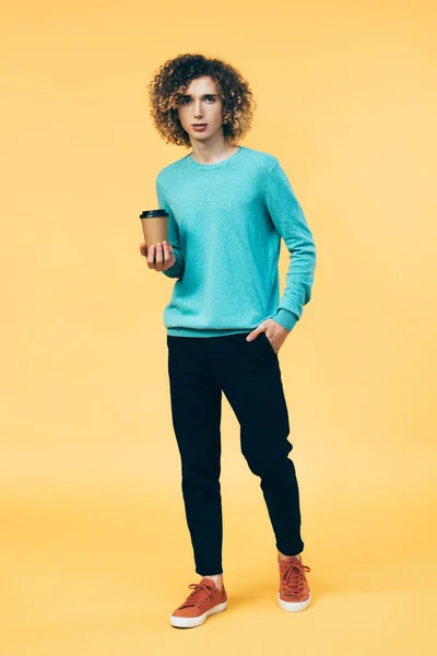 Lockiger Teenager mit der Hand in der Tasche, Kaffee in Pappbecher isoliert auf gelb haltend — Stockfoto