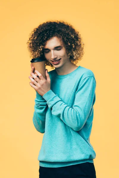 Smiling curly teenager holding coffee in paper cup isolated on yellow — Stock Photo