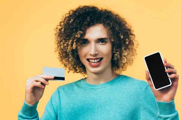 Smiling curly teenager holding smartphone and credit card isolated on yellow — Stock Photo