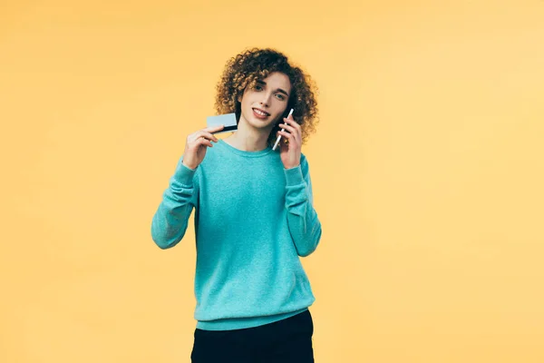 Adolescente rizado hablando en el teléfono inteligente y la celebración de la tarjeta de crédito aislado en amarillo - foto de stock