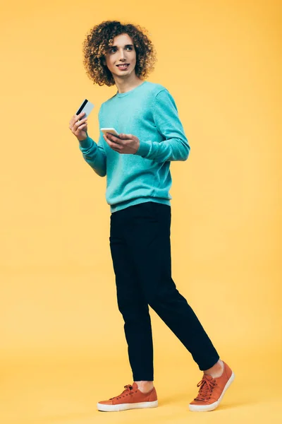 Visão de comprimento total do adolescente encaracolado sorridente segurando smartphone e cartão de crédito enquanto caminha no amarelo — Fotografia de Stock