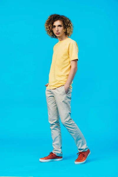 Вид в полный рост кудрявого подростка с руками в карманах, изолированных на голубом фоне — стоковое фото