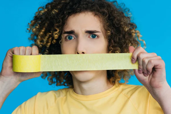 Worried curly teenager holding duct tape near mouth isolated on blue — Stock Photo