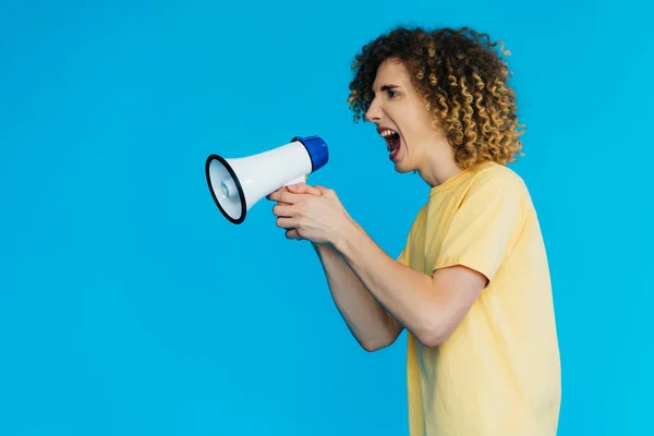 Side view of angry curly teenager screaming in loudspeaker isolated on blue — Stock Photo