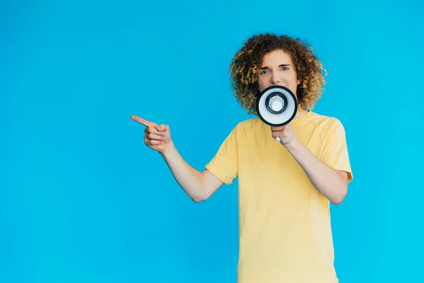 Angry curly teenager screaming in loudspeaker and pointing with finger isolated on blue — Stock Photo