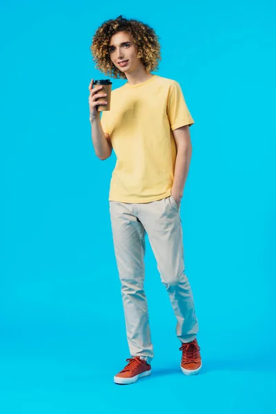 Curly teenager with hand in pocket holding coffee to go isolated on blue — Stock Photo