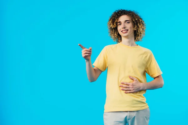 Sonriente rizado adolescente tocando el estómago y señalando con el dedo de distancia aislado en azul - foto de stock