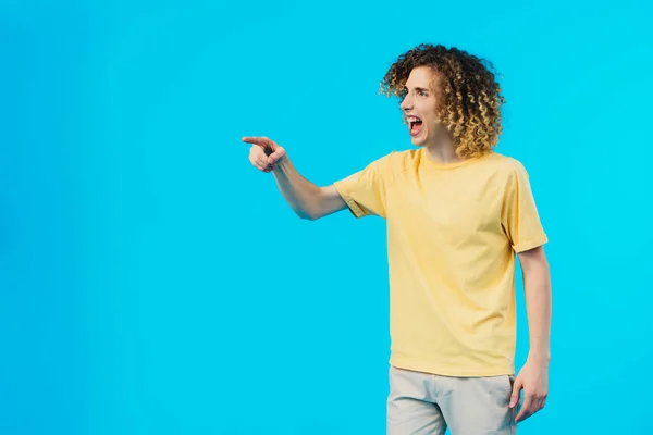 Angry curly teenager yelling and pointing with finger isolated on blue — Stock Photo