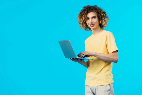 Sonriente estudiante rizado sosteniendo portátil con pantalla en blanco aislado en azul - foto de stock