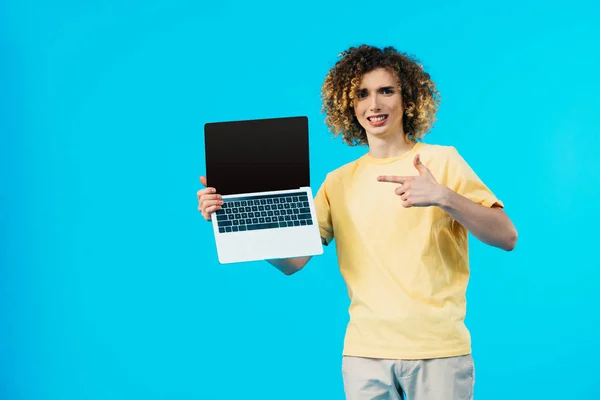 Studente riccio sorridente che punta al computer portatile con schermo vuoto isolato sul blu — Foto stock
