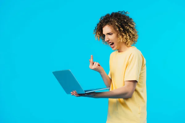 Arrabbiato studente riccio mostrando dito medio al computer portatile isolato su blu — Foto stock
