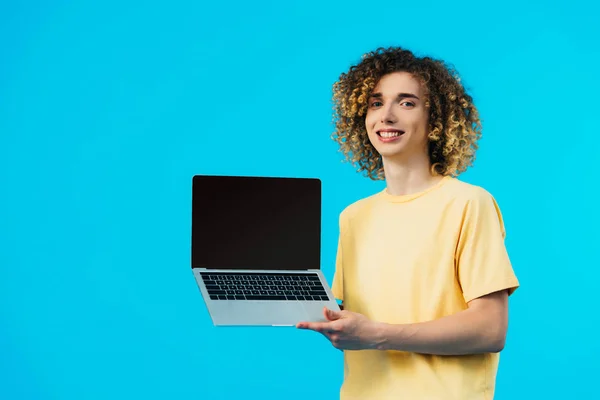 Smiling curly student holding laptop with blank screen isolated on blue — Stock Photo