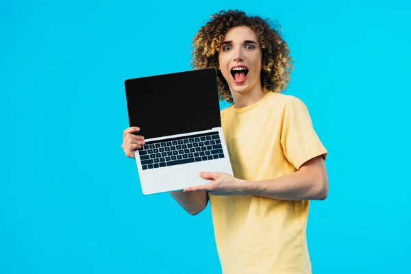 Збуджений кучерявий студент тримає ноутбук з порожнім екраном ізольовано на синьому — стокове фото