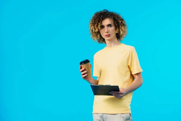 Étudiant bouclé tenant presse-papiers et café pour aller isolé sur bleu — Photo de stock