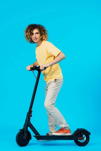 Vue latérale de l'adolescent bouclé équitation scooter électrique sur fond bleu — Photo de stock