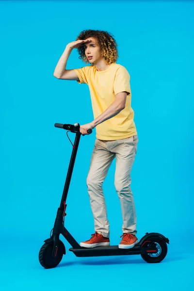 Vue pleine longueur de scooter électrique d'équitation adolescent bouclé et regardant loin sur fond bleu — Photo de stock