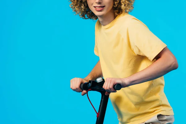 Recortado vista de sonriente rizado adolescente montar scooter eléctrico aislado en azul - foto de stock