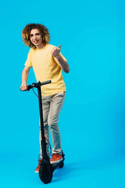 Sonriente rizado adolescente a caballo scooter eléctrico y mostrando el pulgar hacia arriba en azul - foto de stock