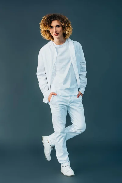 Vista full length di adolescente riccio sorridente in total white outfit con le mani in tasche isolate sul verde — Foto stock