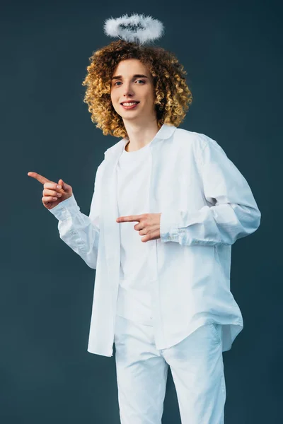 Sorridente adolescente riccio in costume da angelo con alone sopra la testa che punta con le dita isolate sul verde — Foto stock