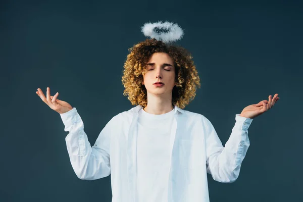 Curly teenager in angel costume with halo above head, closed eyes and outstretched hands isolated on green — Stock Photo