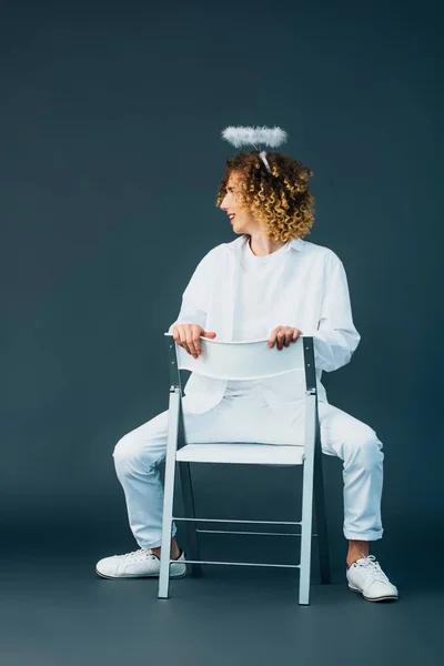 Smiling curly teenager in angel costume with halo above head sitting on chair on green — Stock Photo