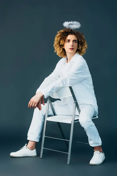 Dreamy curly teenager in angel costume with halo above head sitting on chair on green — Stock Photo