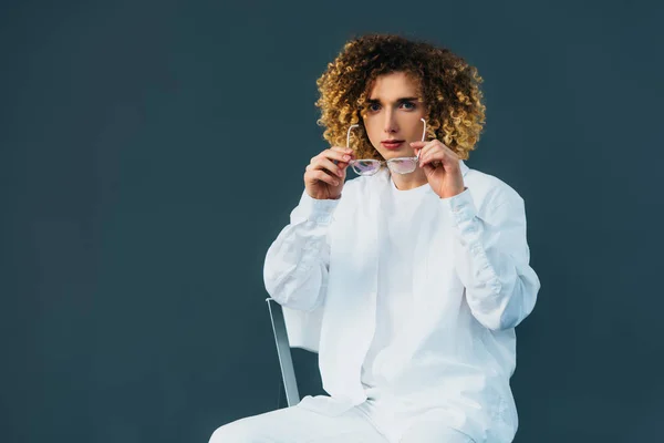 Stylish curly teenager in total white outfit putting on glasses while sitting on chair isolated on green — Stock Photo