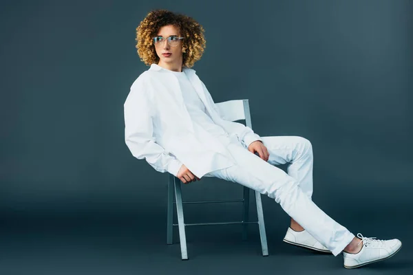 Stylischer lockiger Teenager in komplett weißem Outfit und Brille, der wegschaut, während er auf einem Stuhl auf grün sitzt — Stockfoto
