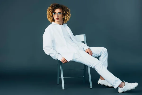 Elegante rizado adolescente en traje blanco total y gafas mirando hacia otro lado mientras está sentado en la silla en verde - foto de stock