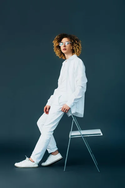 Stylish curly teenager in total white outfit and glasses looking away near chair on green — Stock Photo