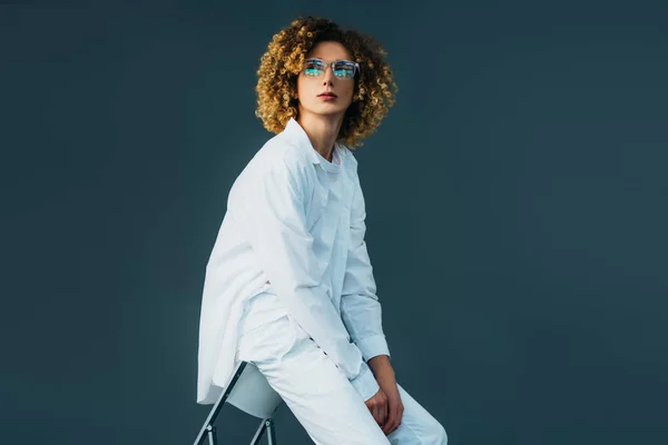 Stylish curly teenager in total white outfit and glasses looking away while sitting on chair isolated on green — Stock Photo