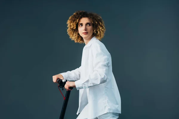 Stylish curly teenager in total white outfit riding electric scooter isolated on green — Stock Photo