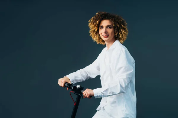Sourire élégant adolescent bouclé en tenue blanche totale équitation scooter électrique isolé sur vert — Photo de stock