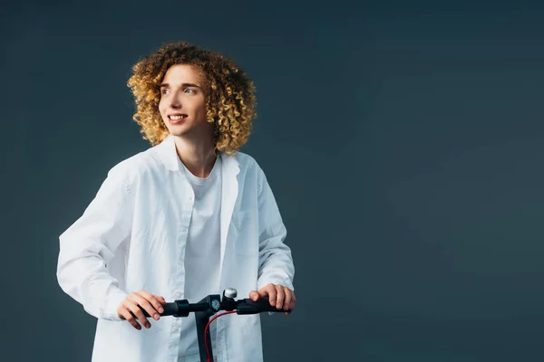 Улыбаясь стильный кудрявый подросток в общей белой одежде езда на электрическом скутере и глядя в сторону изолированы на зеленый — стоковое фото