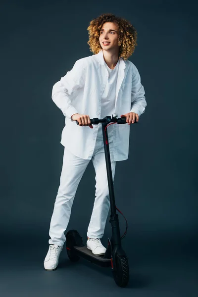 Sourire élégant adolescent bouclé en tenue blanche totale équitation scooter électrique sur vert — Photo de stock