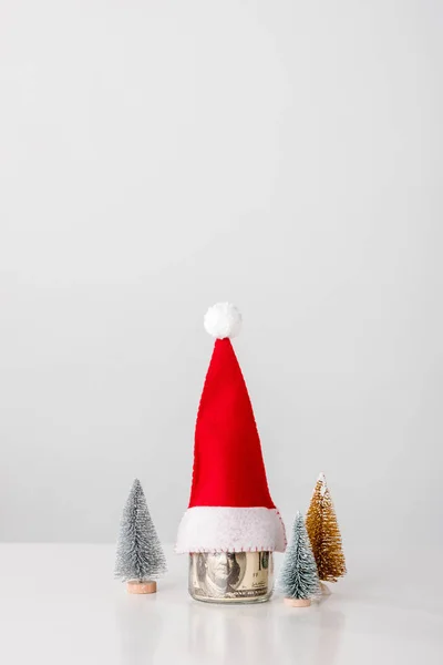 Dekorative und kleine Kiefern in der Nähe von Glas mit Dollarnoten mit Weihnachtsmütze auf weißem Hintergrund — Stockfoto