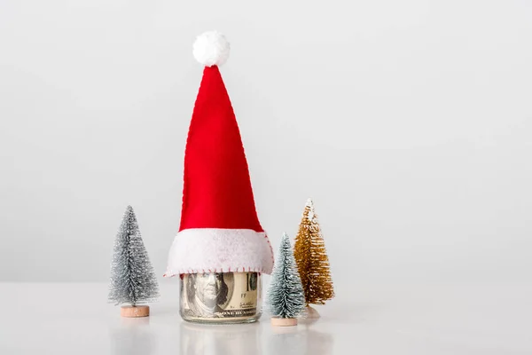 Petits pins décoratifs près du bocal en verre avec billets en dollars avec chapeau de Père Noël sur blanc — Photo de stock